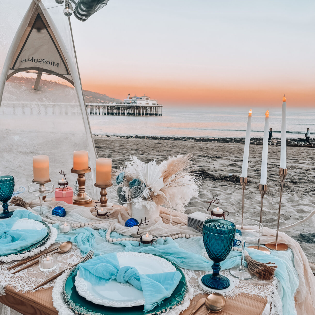 Pop-up picnic scene featuring flickering candles, an elegant dried flower arrangement, bubble tent and light blue drapes, with the Malibu Pier, ocean waves, and the sunset the backdrop.
